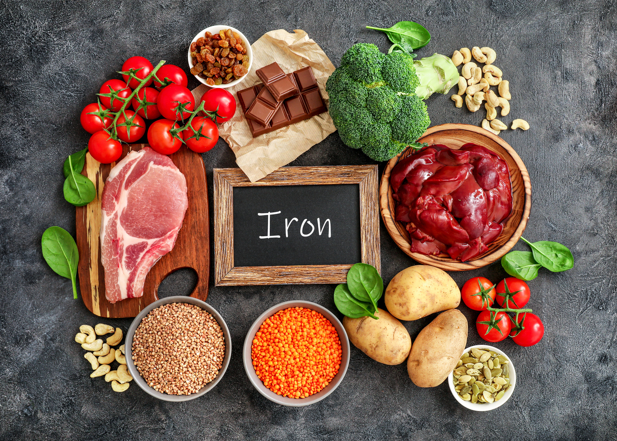 Iron rich foods surrounding a chalkboard including liver, beef, raisins, keshew, buckwheat, spinach, tomatoes, potatoes, dark chocolate, pumpkin seeds, lentil, and broccoli. and potatoes