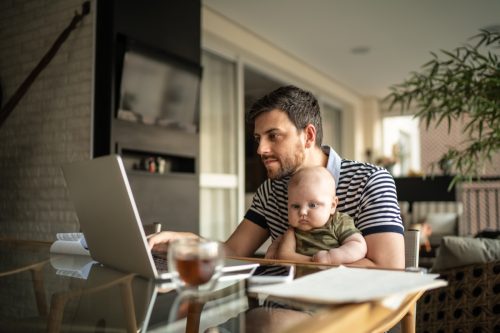 Ein Mann, der seinen Sohn trägt und zu Hause mit seinem Laptop arbeitet