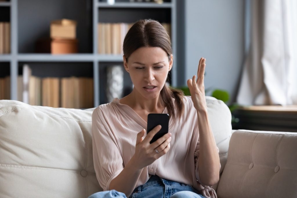 Confused angry woman having problem with phone, sitting on couch at home