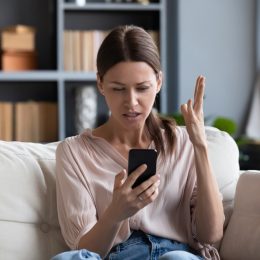 Confused angry woman having problem with phone, sitting on couch at home, unhappy young female looking at screen, dissatisfied by discharged or broken smartphone, reading bad news in message