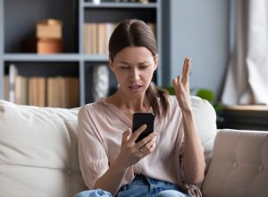 Confused angry woman having problem with phone, sitting on couch at home, unhappy young female looking at screen, dissatisfied by discharged or broken smartphone, reading bad news in message