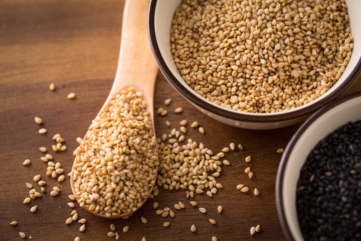 white bowl of sesame seeds next to a wooden spoon full of sesame seeds on wooden counter