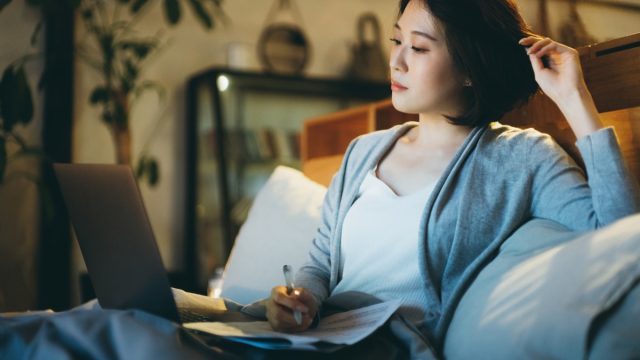 woman sitting on the bed, working from home using laptop and handling paperworks