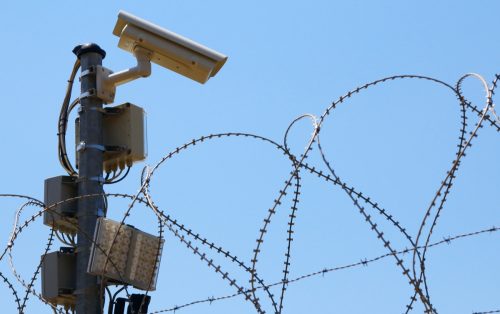 "Surveillance camera, barbed wire fence and searchlight against blue sky."