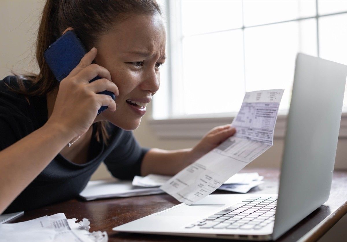 upset woman disputing a charge on the phone while holding a bill and looking at laptop