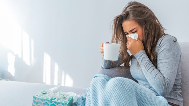 A woman sitting on the couch while sick and blowing her nose into a tissue while holding a mug