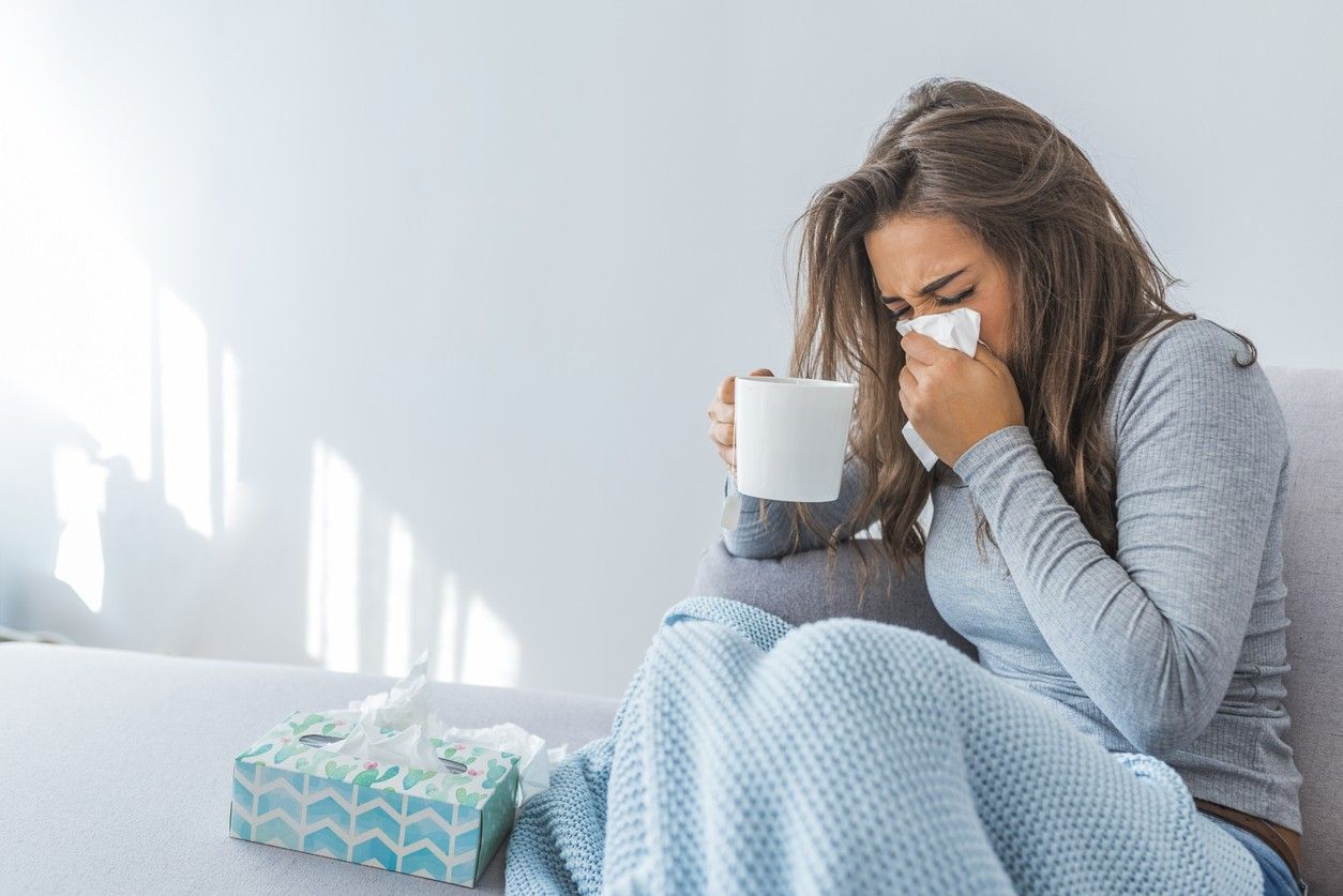 A woman sitting on the couch while sick and blowing her nose into a tissue while holding a mug