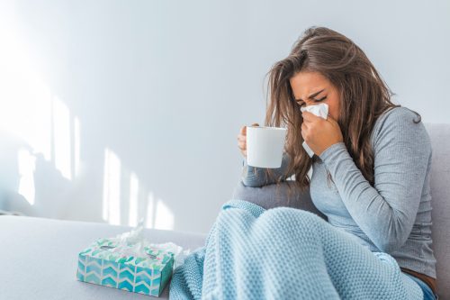 A woman sitting on the couch while sick and blowing her nose into a tissue while holding a mug