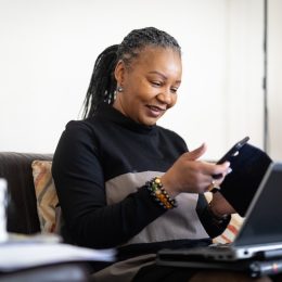 middle-aged woman wearing a black turtleneck with her hair in dreadlocks looking at phone and computer on couch