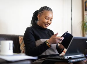 middle-aged woman wearing a black turtleneck with her hair in dreadlocks looking at phone and computer on couch
