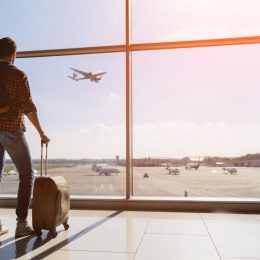 male tourist is standing in airport and looking at aircraft flight through window. He is holding tickets and suitcase. Sunset