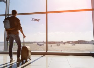 male tourist is standing in airport and looking at aircraft flight through window. He is holding tickets and suitcase. Sunset