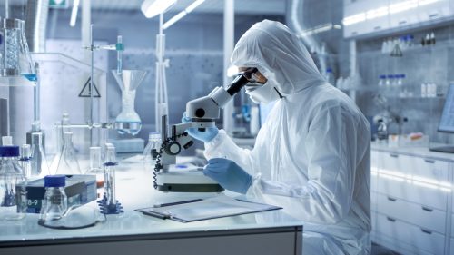 A scientist completing a study in a lab looking into a microscope while wearing full protective gear
