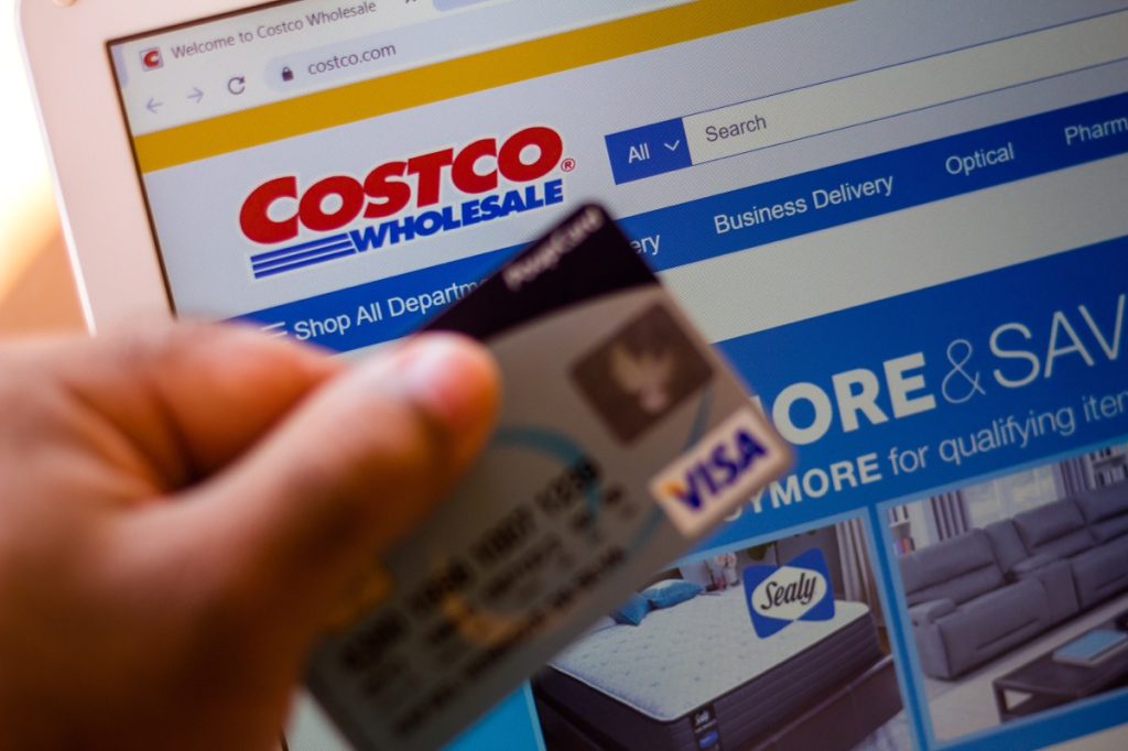 The Costco Wholesale Corporation website is shown on a laptop in the background and a person holds a bank card