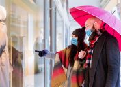 couple with an umbrella and winter clothes is talking and smiling while doing shopping in the city in a rainy day. side view of a couple walking past a shop window and looking in for sales.