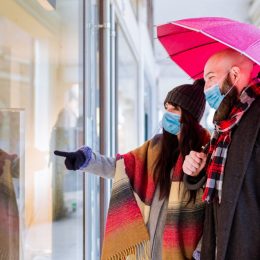 couple with an umbrella and winter clothes is talking and smiling while doing shopping in the city in a rainy day. side view of a couple walking past a shop window and looking in for sales.
