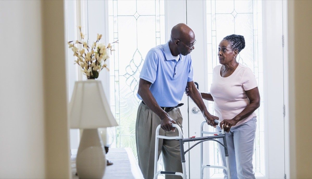 couple inside their home, at the front door. The man is using a mobility walker and his wife is helping him