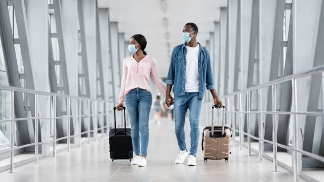 Couple Wearing Protective Masks Walking With Suitcases At Airport Terminal, Ready For Post-Quarantine Flight, Copy Space