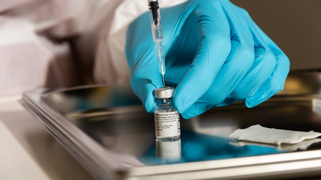 A gloved hand filling a syringe from a Pfizer vaccine vial