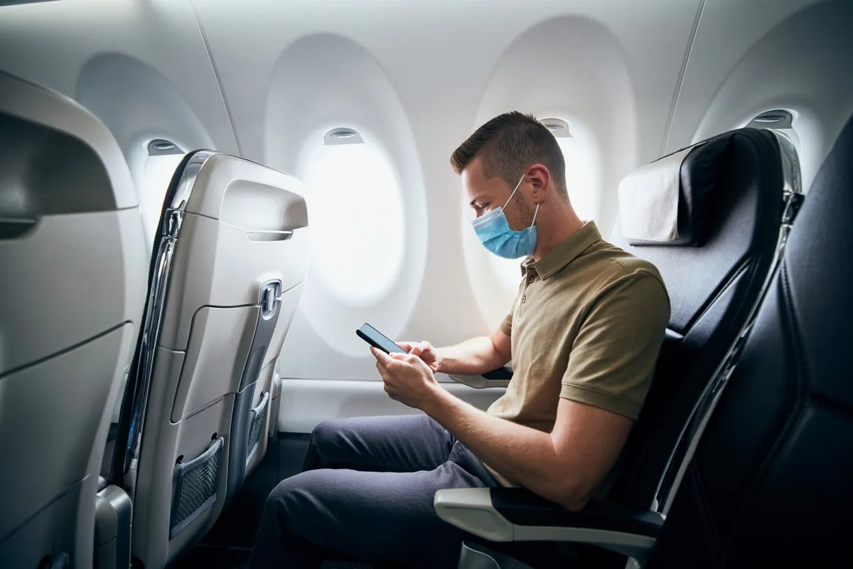 Ein Mann trägt eine Gesichtsmaske und benutzt während des Fluges ein Telefon im Flugzeug.  Natürlich neue Themen, Corona-Virus und Personenschutz.