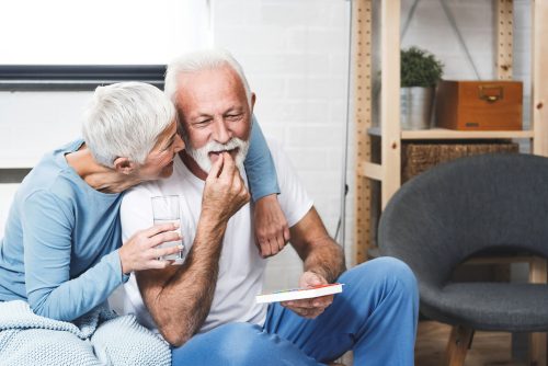 A senior man being hugged by his wife while taking a pill and sitting on the couch