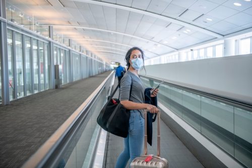A young female traveler walking escalator at the airport. She has her carry on suit case and a bag on her. She is wearing a face mask to prevent the spread of germs.