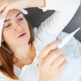A young woman lying on the couch while feeling sick, taking her temperature and holding a compress to her forehead