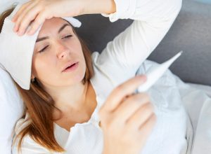 A young woman lying on the couch while feeling sick, taking her temperature and holding a compress to her forehead