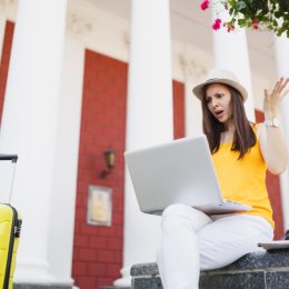 A young woman traveler sitting with her suitcase and looking angrily at her laptop