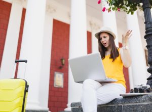 A young woman traveler sitting with her suitcase and looking angrily at her laptop