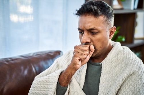 Shot of a man coughing while recovering from an illness on the sofa at home