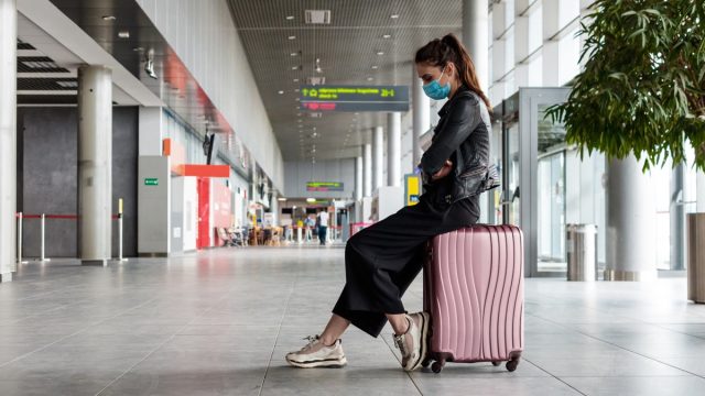 People traveling by plane during COVID 19. Young woman wearing N95 face masks, sitting on luggage in airport terminal.