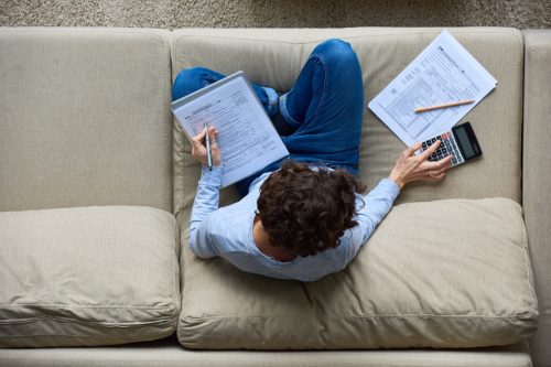 Directly above view of concentrated accountant using calculator while preparing tax forms sitting on sofa