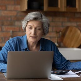 Smiling mature woman using laptop, online baking service, checking financial documents at home, senior grey haired female sitting at table with domestic bills and calculator, accounting