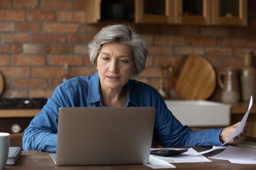 Smiling mature woman using laptop, online baking service, checking financial documents at home, senior grey haired female sitting at table with domestic bills and calculator, accounting