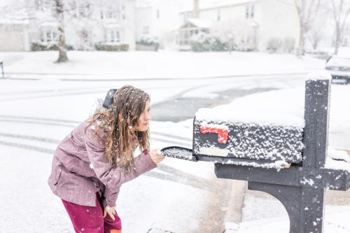 Mujer joven revisando el correo en la carretera del vecindario con suelo cubierto de nieve durante la tormenta blanca, copos de nieve cayendo en los suburbios de Virginia, casas unifamiliares en el buzón