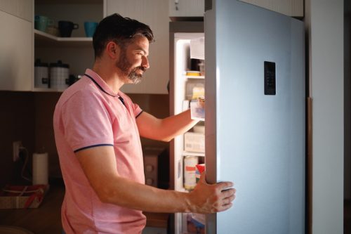 Close-up side view of man searching refrigerator for food, his face is lit by refrigerator light