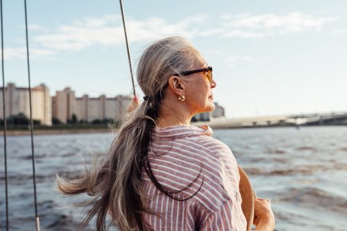 Side view of mature woman with long gray hair wearing sunglasses and looking to the distance while standing on a yacht on the water