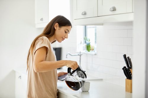Side view of young woman pouring coffee in cup. Beautiful female is standing at kitchen counter. She is in casual at home.