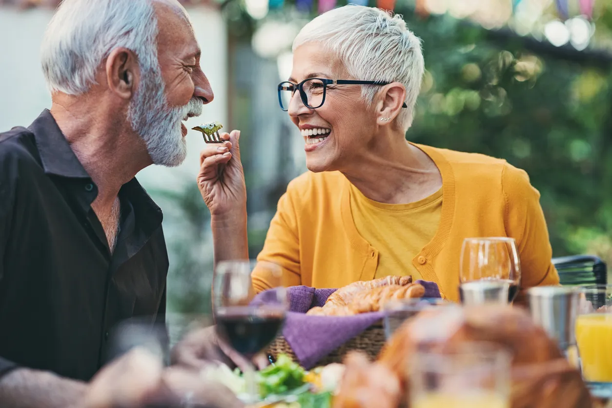 A senior woman playfully feeding a man from a fork while eating outside at a dinner party