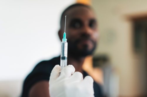 ภาพระยะใกล้ของแพทย์ที่ฉีดวัคซีนให้ชายหนุ่มที่บ้านเพื่อฉีดวัคซีนป้องกัน covid-19  หมอหญิงถือเข็มฉีดยาเพื่อเตรียมวัคซีนโควิด-19