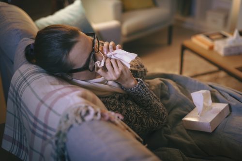 Tânără care își sufla nasul în timp ce este bolnavă pe canapea din cauza COVID sau a gripei