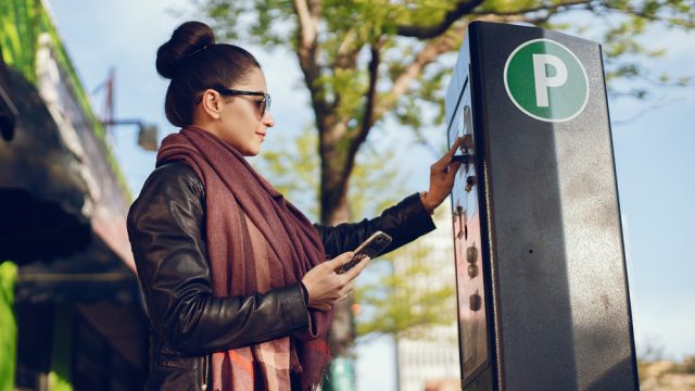 A young woman paying at a public parking meter terminal while holding her phone