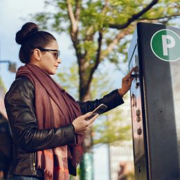 A young woman paying at a public parking meter terminal while holding her phone