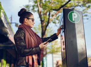A young woman paying at a public parking meter terminal while holding her phone