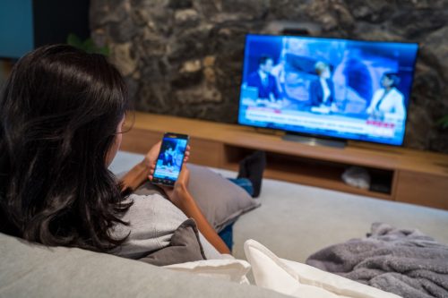 Rear view of young woman watching news on television and smart phone while sitting on sofa in living room.