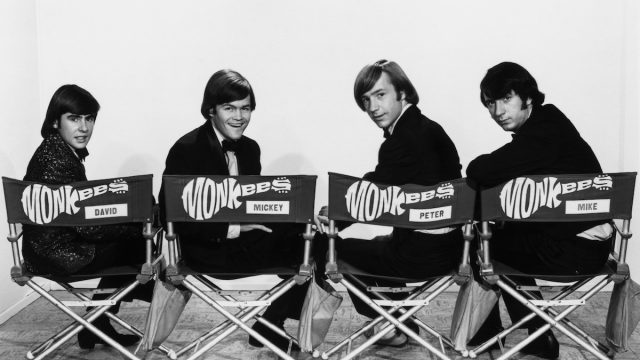 The Monkees sitting in chairs with their names on them and looking over their shoulders