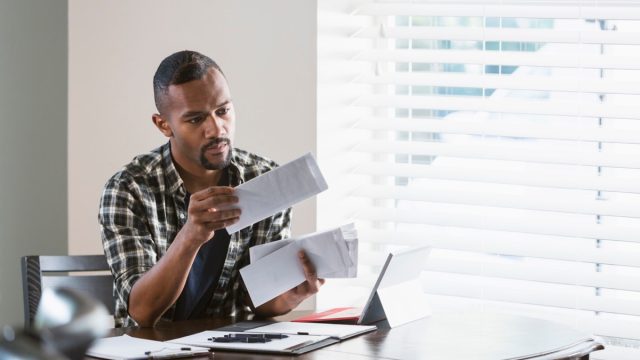 man sitting at a desk by a window at home, paying bills. He is looking through a stack of envelopes and has his laptop computer in front of him.