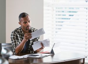 man sitting at a desk by a window at home, paying bills. He is looking through a stack of envelopes and has his laptop computer in front of him.