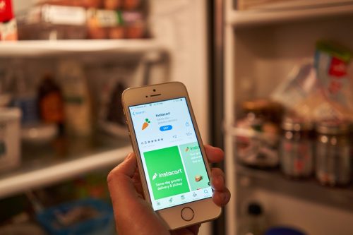 Жена проверява мобилното приложение Instacart на телефона си, докато отваря хладилника.  Instacart предлага услуга за доставка и получаване на хранителни стоки в същия ден